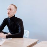 Young man in a black turtleneck writing at a desk, contemplating ideas.